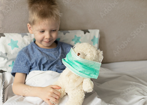 A sick sad child with a temperature and a headache lies in bed with a toy teddy bear. Flu colds disease virus bacterium.