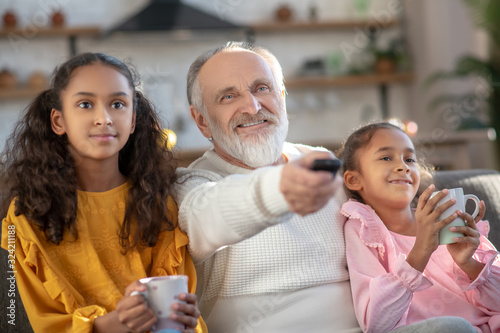 Two dark-skinned girls sitting next to their granddad and watching tv