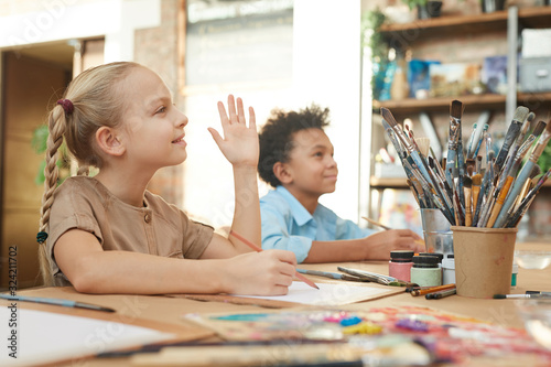 Little girl raising her hand while sitting at the table and painting with boy in the background during art lesson