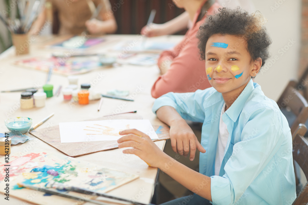 Portrait of African boy looking at camera while sitting at the table with paper and paints at art lesson Portrait of African boy looking at camera while sitting at the table with paper and paints at art lesson
