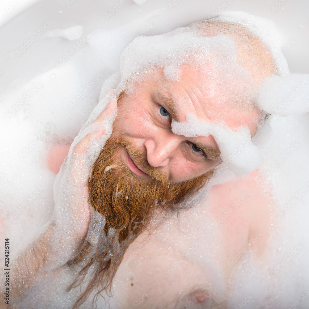 Portrait of a bald man with a long red beard takes a bath with foam. Top view on a cheerful funny guy in soapy water. Portrait of a bald man with a long red beard takes a bath with foam. Top view on a cheerful funny guy in soapy water.