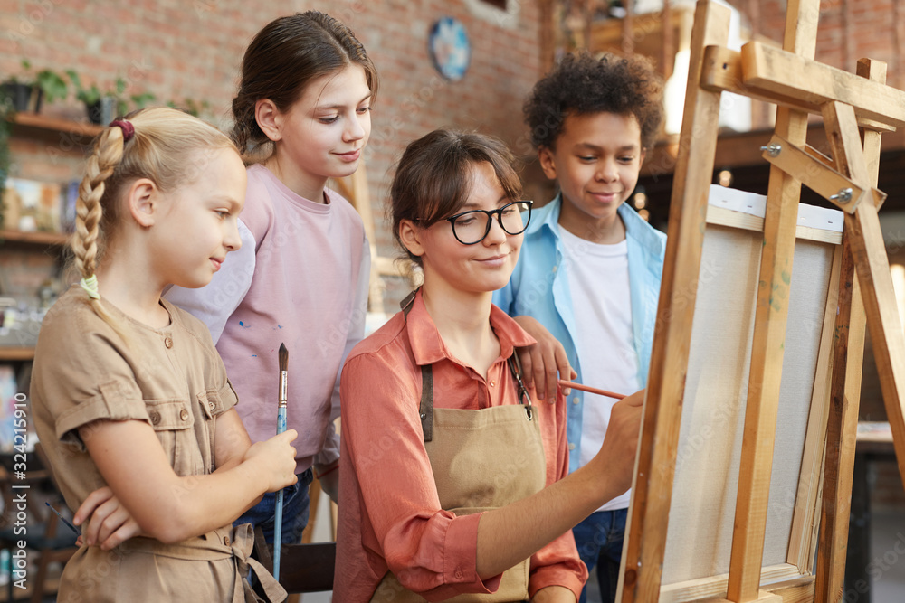 Young woman painting on easel and group of children watching for the process at art studio Young woman painting on easel and group of children watching for the process at art studio