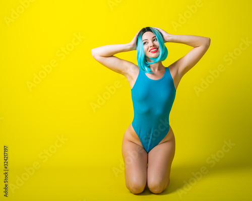 Portrait of a blue hair woman in a wig and a swimsuit on a yellow background. Fashion Girl in a monokini with artificial hair posing in the studio. © Михаил Решетников