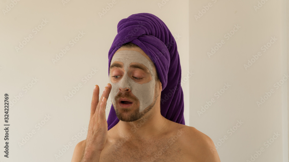 Funny handsome young man with purple towel on his head and face cosmetic mask after bath .Man wears facial clay mask enjoys beauty tratments.Funny content for Womens Day, spa or Valentine day. Funny handsome young man with purple towel on his head and face cosmetic mask after bath .Man wears facial clay mask enjoys beauty tratments.Funny content for Womens Day, spa or Valentine day.