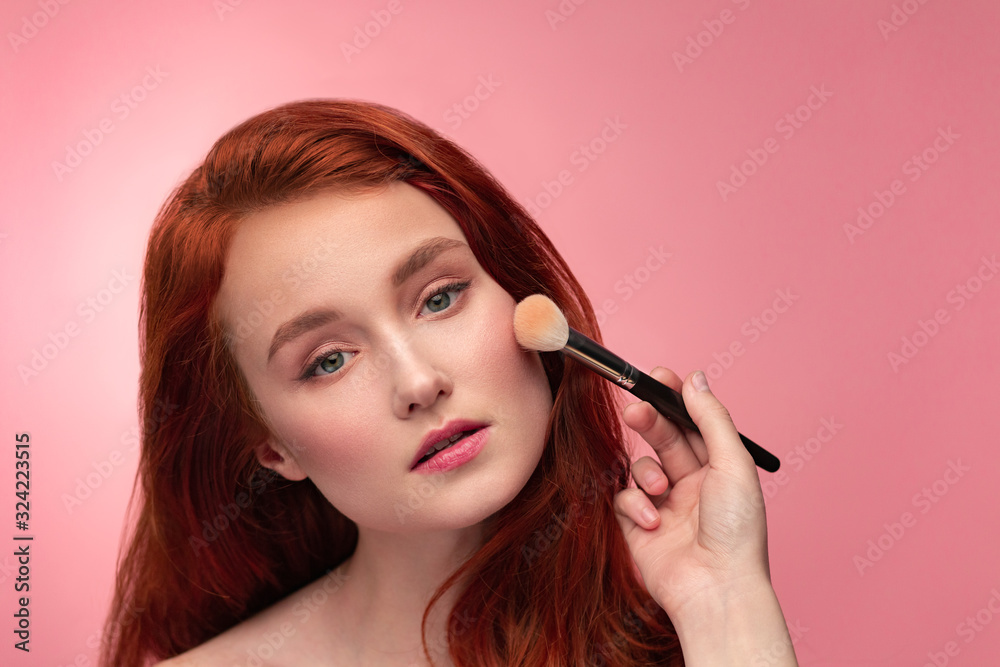 Fototapeta premium Makeup artist applies powder. Hand of make-up master, painting cheeks of young redhead girl. Portrait beautiful healthy girl with pure clean skin and natural make-up. Beauty Concept. Pink background.