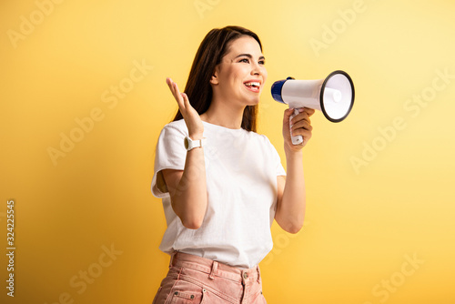 happy girl speaking in megaphone while standing with open arm and looking away on yellow background