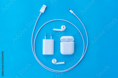 Top view flat lay plastic white wireless headphones for smartphone, charging case, Lighning USB, adapter on a pastel blue background. Copy space.