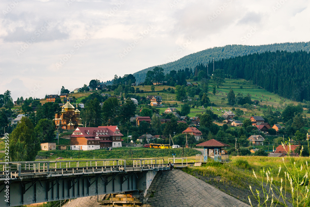 Ukrainian village in the Carpathians in sunny evening. Beautiful landscape during the evening light. Summer country side view. Authentic abandoned railway with traditional houses, church, hills.