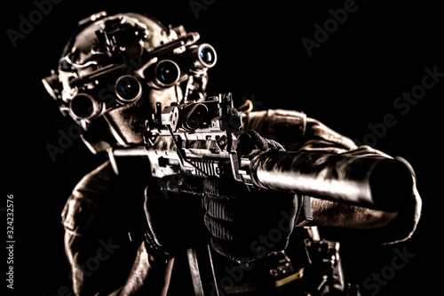 Army special operations soldier, commando fighter in full tactical ammunition, helmet with radio headset and night vision device, aiming short barrel assault rifle in darkness, low key studio shoot