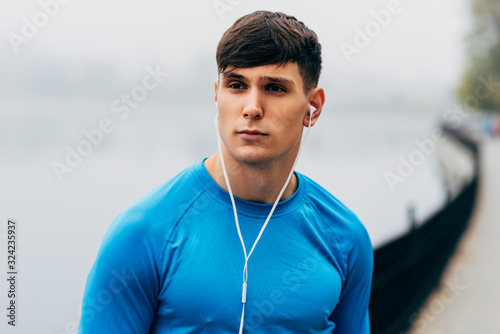 Close-up portrait of young runner man posing during jogging in the morning on the fog lake background. Fitness male exercising in the park and listenting the music on earphones. photo