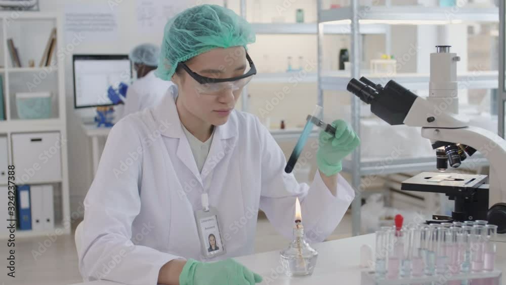 Medium shot of female Asian virologist wearing safety goggles and gloves sitting at desktop in laboratory, holding glass-tube using pincer and heating it on fire Medium shot of female Asian virologist wearing safety goggles and gloves sitting at desktop in laboratory, holding glass-tube using pincer and heating it on fire