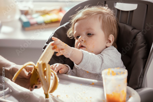  Baby in the child dining chair eat healthy  vegetable food. Child eat  pumpkin and carrot puree with banana. Mother s hand. Dirty face of happy kid. Portrait of a baby eating with a stained face.