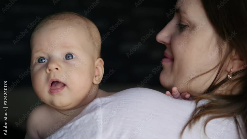 Lovely baby leaning on mother’s shoulder and looking around with big blue eyes. A woman holds a small baby in her arms. Lovely baby leaning on mother’s shoulder and looking around with big blue eyes. A woman holds a small baby in her arms.