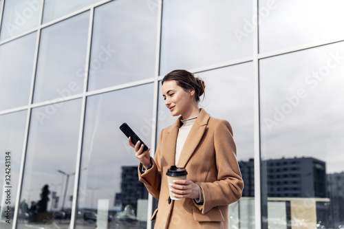 Smiling curly woman wearing trendy sunglasses walks down the central city street and uses her phone. Pretty summer woman in brown jacket walks down the street looking at her mobile phone