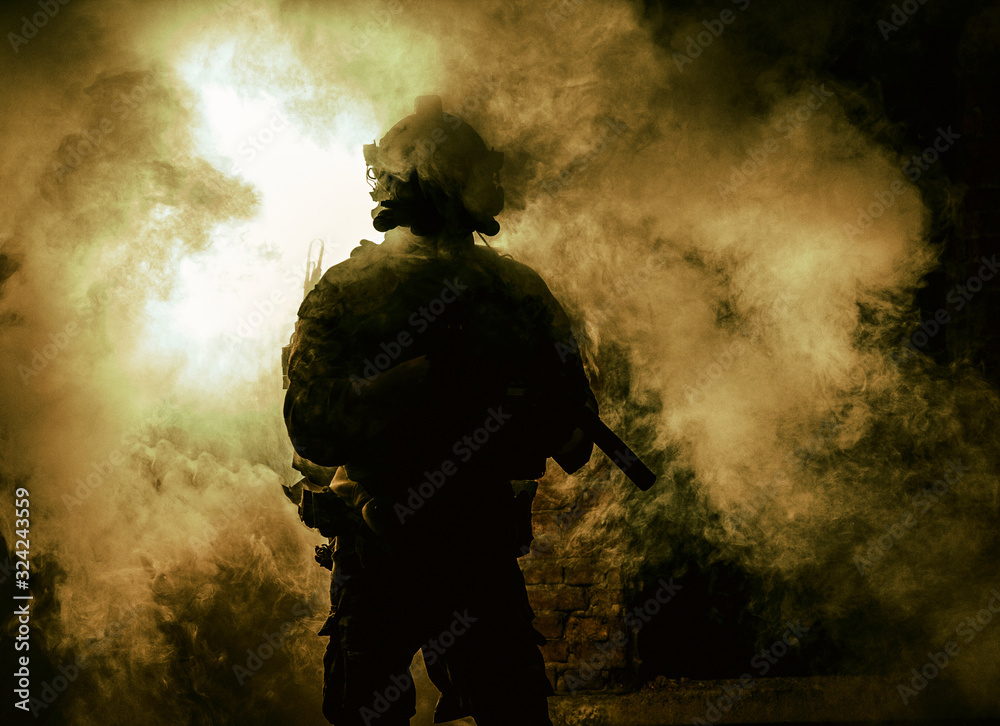 Fototapeta premium Silhouette of modern infantry soldier, elite army fighter in tactical ammunition and helmet, standing with assault service rifle in hands on background of fiery explode. Burning fire of war conflict