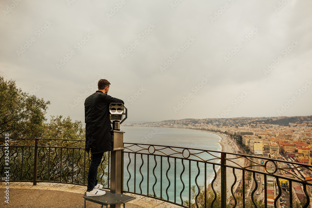 A guy stands on the viewing platform and looks through binoculars on a beach and a waterfront of Nice, France. Panorama of Nice city, Cote d'Azur, French riviera, Promenade des Angles.