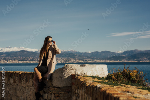 Lovely elegant girl in beige coat and black dress with glasses visiting the famous Mediterranean Antib port. Traveling woman on vacation in France. Stylish girl in the port relaxing.