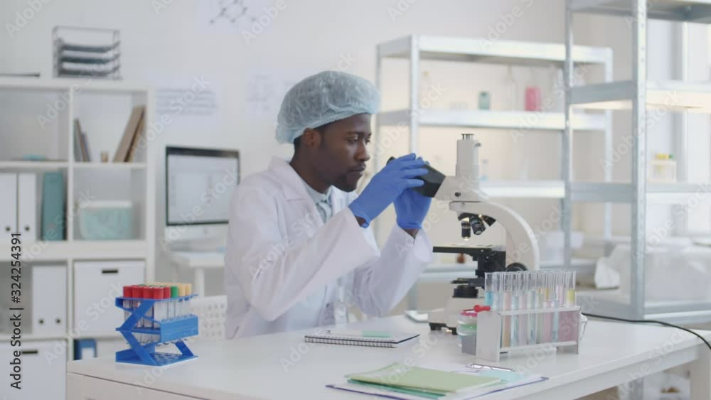 Lockdown of African scientist wearing special uniform sitting down at desk and starting examining sample using microscope Lockdown of African scientist wearing special uniform sitting down at desk and starting examining sample using microscope