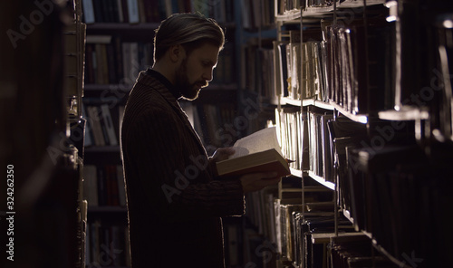Bilde på lerret Man consumed with reading in dark library standing among rows of shelves with bo