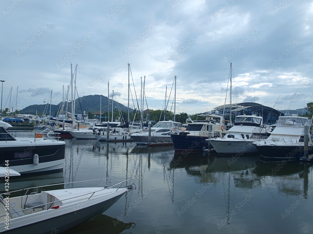 Yachts in the bay, reflection from water. Sunset dark cloudy sky, sailing yachts and powerboats moored on the pier of a tropical luxury marine Phuket Boat Lagoon yacht club in the tropical island.