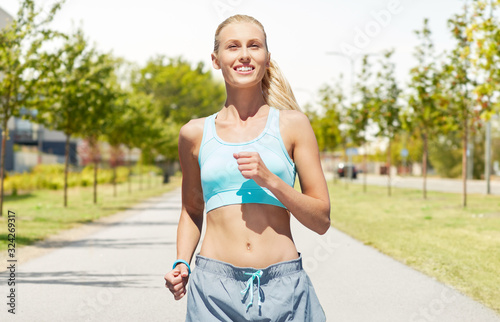 sport, healthy lifestyle and people concept - smiling young woman with fitness tracker running along road on city street background