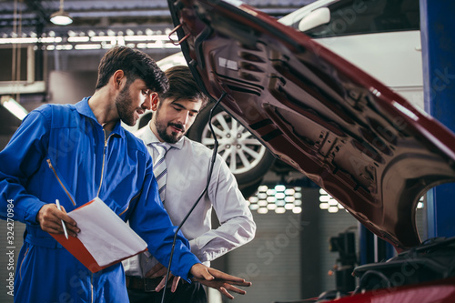 automotive maintenance mechanic young man explain car condition to male customer in garage at auto repair shop, technician pointing needed on vehicle checklist document, after service concept
