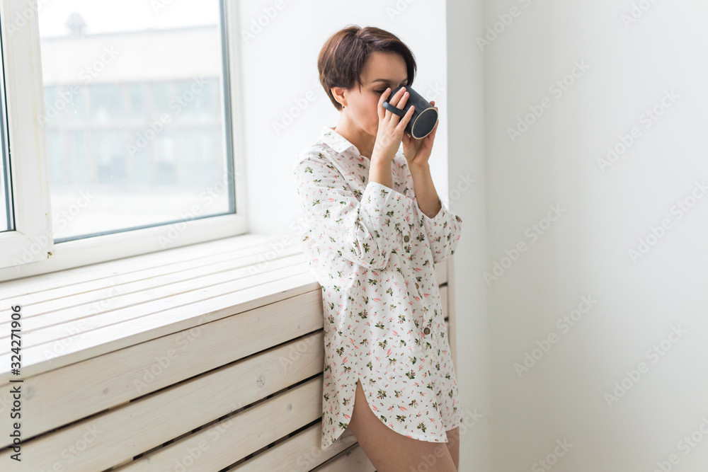 Calm and coziness. Beautiful young woman with cup of tea standing near the window at home Calm and coziness. Beautiful young woman with cup of tea standing near the window at home