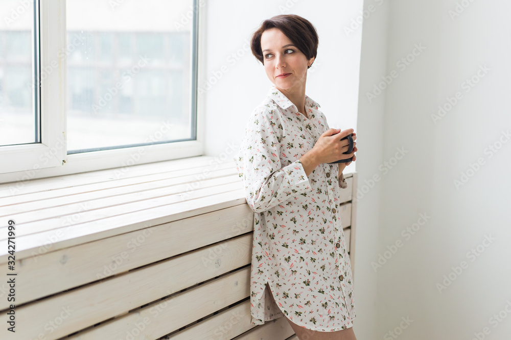 Calm and coziness. Beautiful young woman with cup of tea standing near the window at home Calm and coziness. Beautiful young woman with cup of tea standing near the window at home