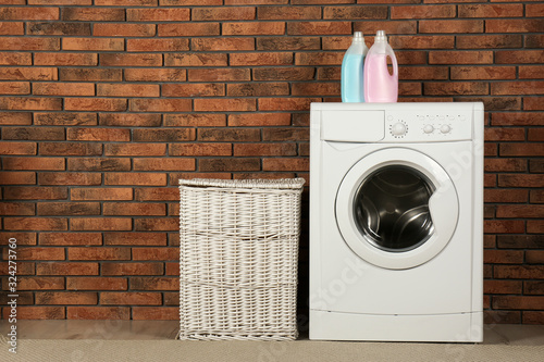 Modern washing machine with detergent and laundry basket near brick wall