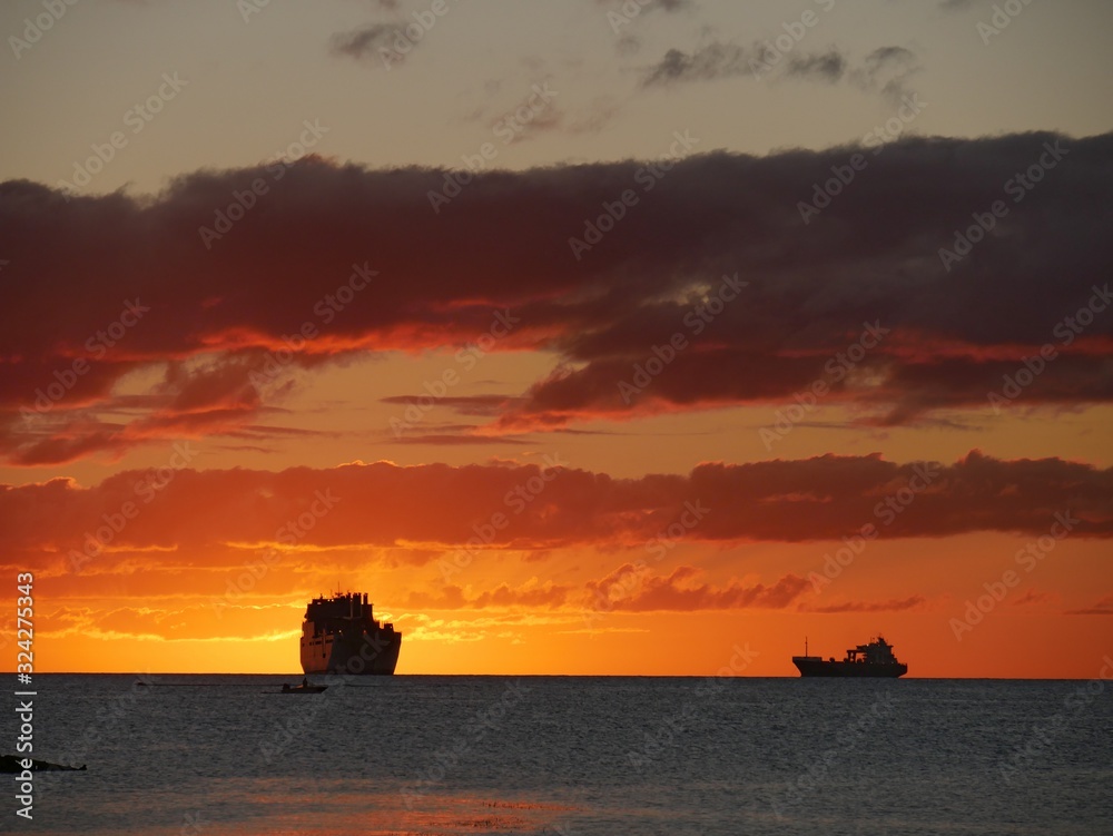 Skies on fire at sunset, with the silhouettes of ships in the distance