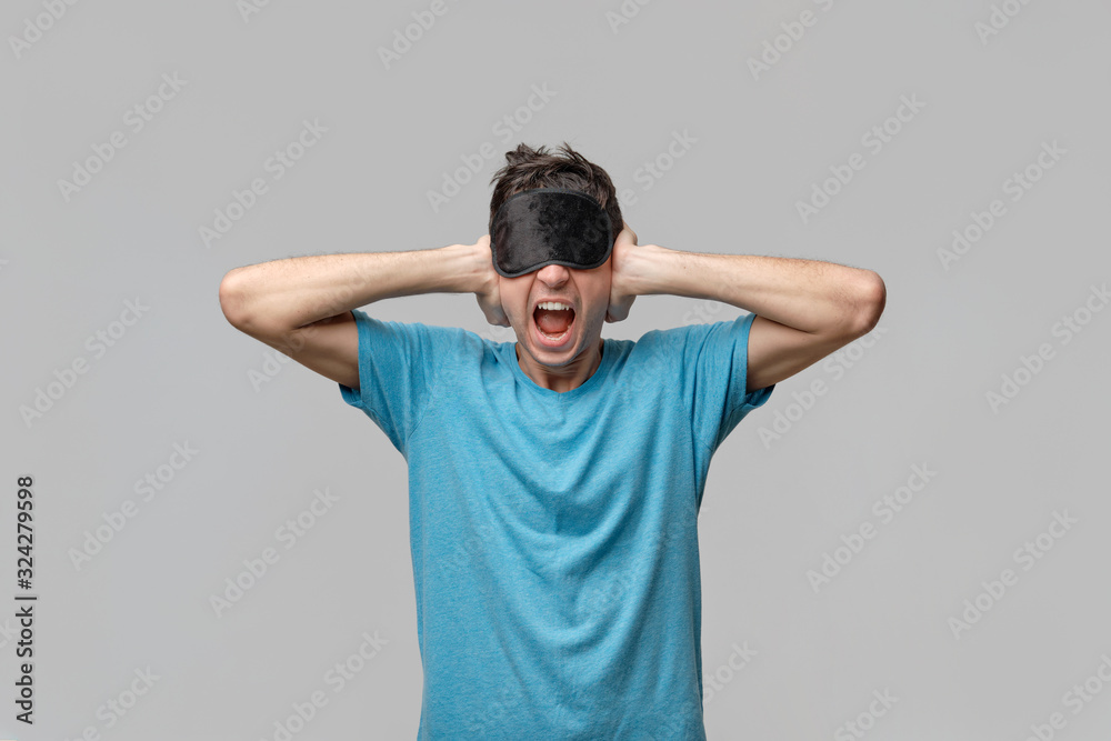 Sleepy man with black sleep mask on a head screaming closing ears isolated over grey background Sleepy man with black sleep mask on a head screaming closing ears isolated over grey background