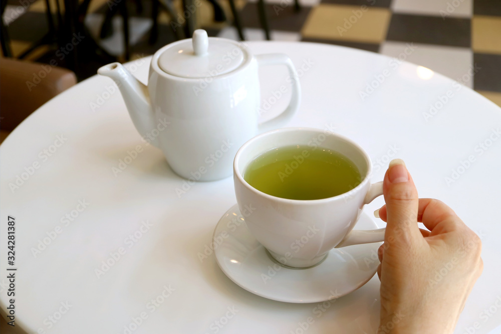 Female's hand holding cup of green tea on a white round table with teapot in background Female's hand holding cup of green tea on a white round table with teapot in background