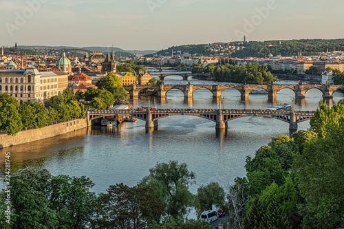 Prague / Czech Republic - May 23 2019: Scenic view of the cityscape, the river Vltava and Charles bridge, Manes and Jirasek bridges across water. Green trees in foreground. Sunny spring evening.