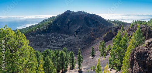 Panoramic landscape with lush green pine trees, colorful volcanoes and lava crater Deseada along path Ruta de los Volcanes, hiking trail at La Palma island, Canary Islands, Spain, Blue sky background