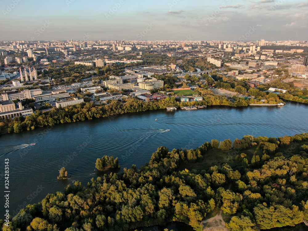 A motor boat floats along the coast of the Moscow River, in the background the city outskirts with buildings and roads, beautiful green nature around. Aerial photography