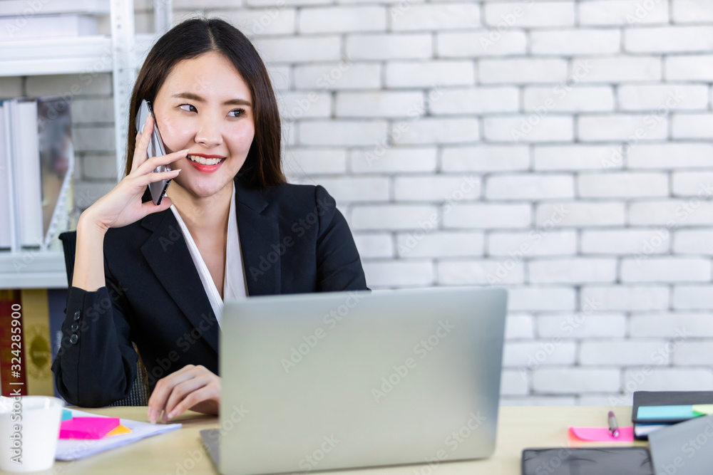 Happy of successful asian young businesswoman hold pick up smartphone working with laptop computer in on White wooden table in white wall office background Happy of successful asian young businesswoman hold pick up smartphone working with laptop computer in on White wooden table in white wall office background