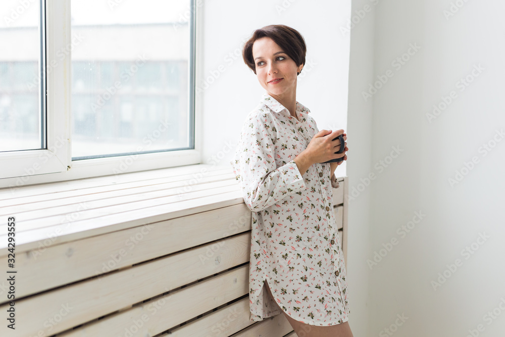 Calm and coziness. Beautiful young woman with cup of tea standing near the window at home Calm and coziness. Beautiful young woman with cup of tea standing near the window at home
