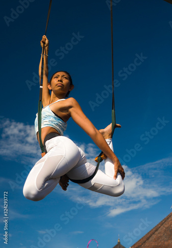outdoors portrait of young happy and athletic Asian Indonesian woman doing aero yoga workout training body balance hanging from swing rope isolated on blue sky