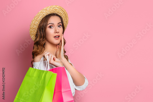 Shocked girl in a straw hat and white dress holds shopping bags in hands isolated on a pink background, copy space. Surprised, excited girl gives gifts , web banner, sale concept