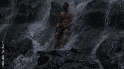 Tracking shot of  young handsome man standing on stone under waterfall on tropical island at dusk