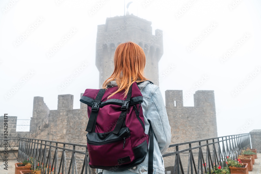 Back view woman with backpack wearing jeans jacket. Fortification on top of the mountain, old castle on background. Journey. Mystical atmosphere, fog, white haze, mist. Second Tower San Marino, Italy