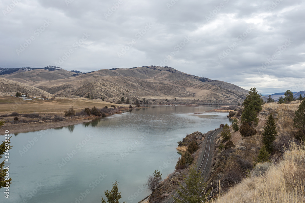 Train track alongside a calm river with large rolling hills in the distance