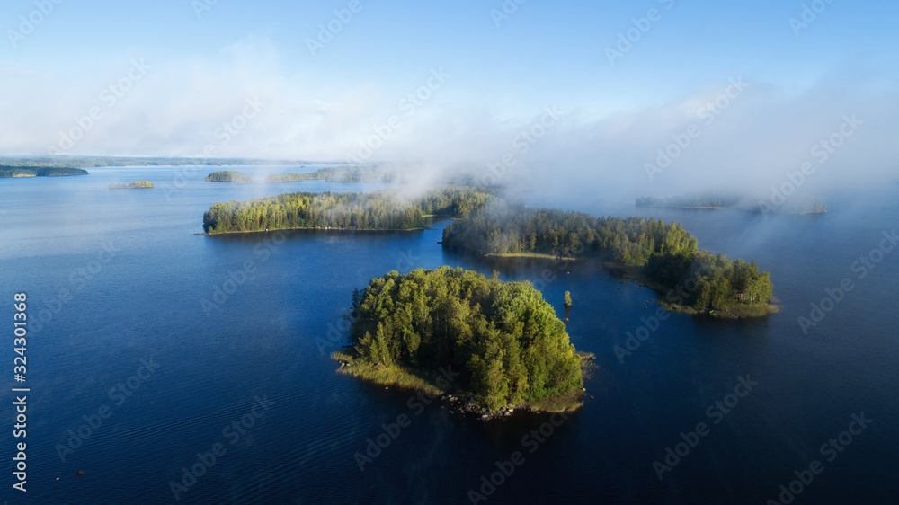 Fototapeta premium Calm lake water with forests and islands. Early misty summer morning with clouds. Finland.