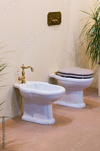 Clean and white toilet and bidet with gold-plated faucet in light bathroom. Beautiful bidet in luxurious bathroom.