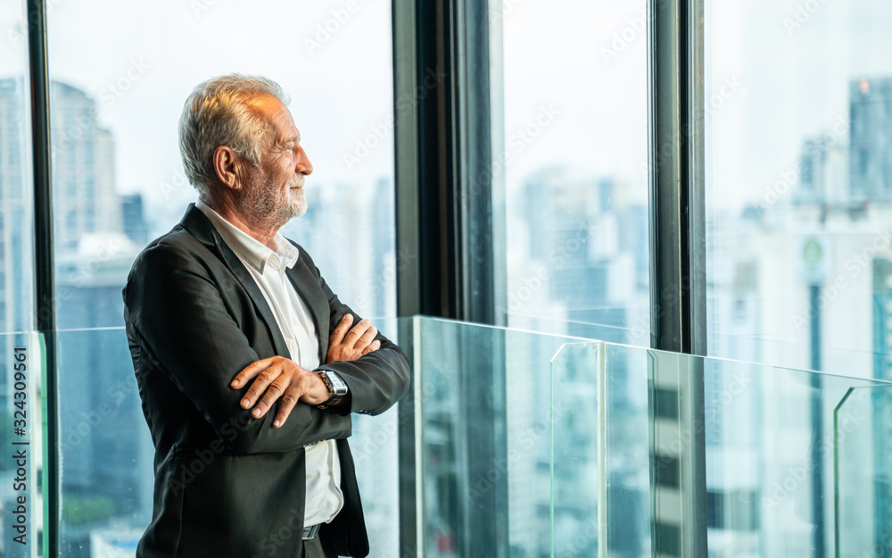 Retirement Man Concept.Portrait of Senior grey-haired businessman in suit standing and and looking through window the cityscape.