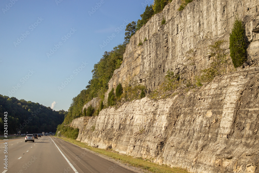 Asphalt road among the high mountains on a sunny summer day. Sheer cliffs overgrown with plants hang over the road. Cars ride on the highway among the mountains