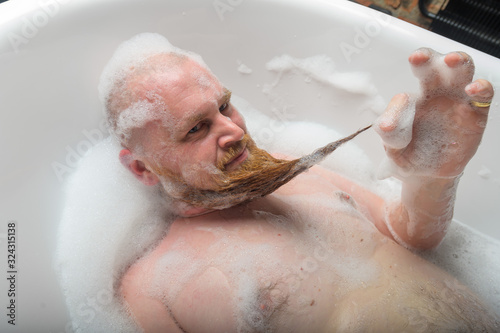 A bald man is fooling around in the foam bath. The guy makes funny hairstyles on his red beard.