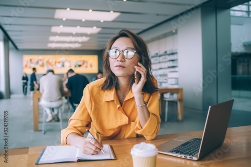 Pensive female freelancer in classic spectacles listening news received with cell phone call during remote work in coworking space, pondering Asian woman communicating with customer service