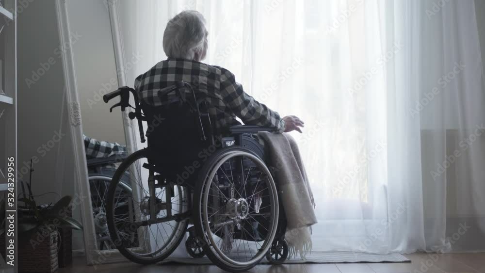 Back view of old Caucasian man sitting in wheelchair in front of big window closed with curtain and thinking. Lonely elderly man spending day alone at home. Oldness, retirement, disabled people.