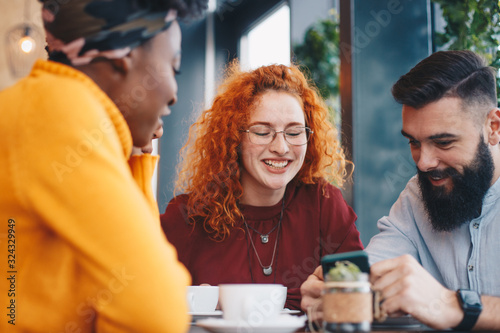 Three friends sitting in a cafe, talking, drinking coffee and looking at the male friend's phone.